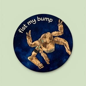 May include: A circular sticker featuring a cartoon character with a brown and tan textured appearance, set against a dark blue, starry background. The text "fist my bump" is written in white around the top of the circle.