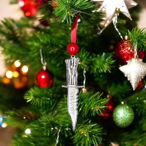 May include: A silver knife ornament with a black and white patterned handle hanging on a green Christmas tree. The ornament is shaped like a knife with a serrated blade.