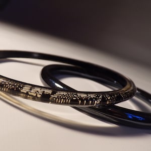 May include: Two black bangle bracelets, one with a clear, patterned design and the other with a solid black finish.