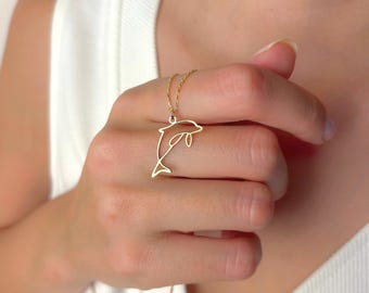 以下が含まれることがあります： 金色のイルカペンダントネックレス。ペンダントはイルカのミニマリストなアウトラインが特徴です。ネックレスは首の周りに着用されています。背景は柔らかく、ニュートラルな色です。