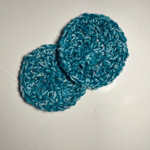 May include: Two crocheted scrubbies in a teal and white color combination. The scrubbies are round and have a textured surface.