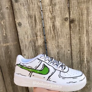 以下が含まれることがあります： 緑と黒の漫画風のデザインが施された白い革のスニーカー。ナイキのスウッシュは緑色で黒で縁取られています。靴の側面に「@labellamarposaco」というテキストがあります。