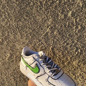 以下が含まれることがあります： 黒のアウトラインと緑のナイキのスウッシュが付いた白いローカットスニーカー。靴は漫画のようなデザインです。