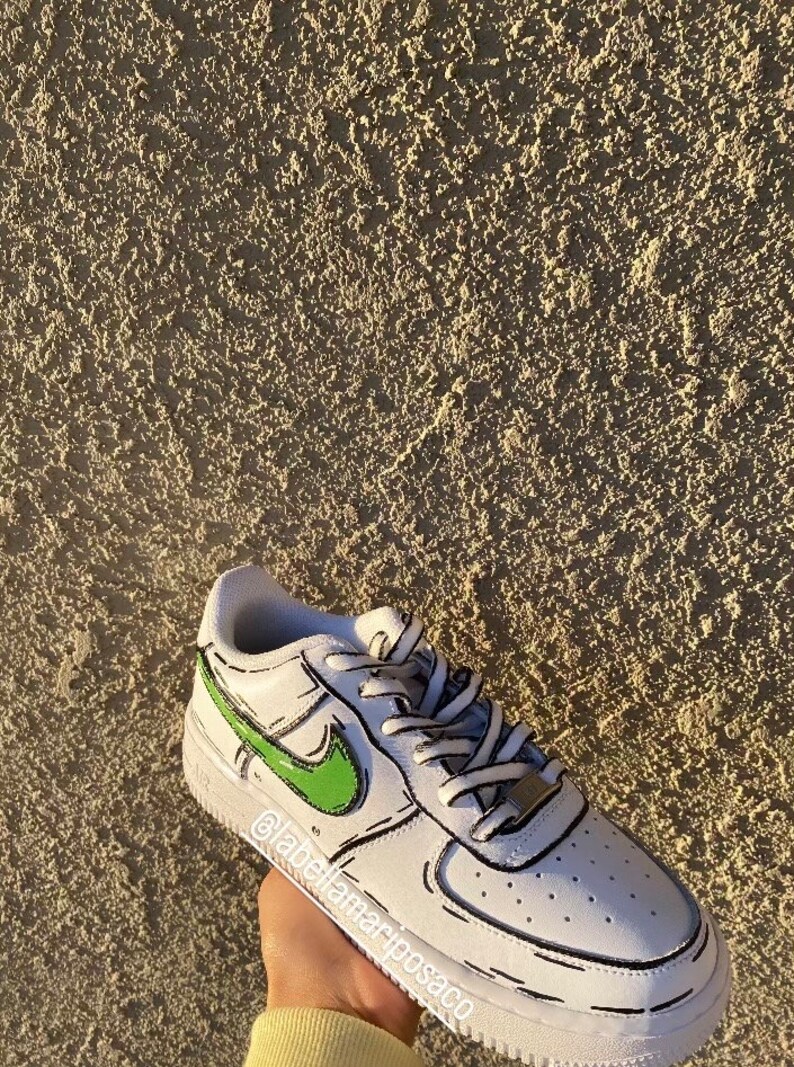 以下が含まれることがあります： 黒のアウトラインと緑のナイキのスウッシュが付いた白いローカットスニーカー。靴は漫画のようなデザインです。