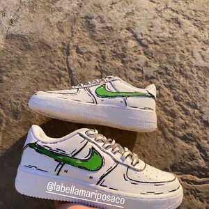 以下が含まれることがあります： 緑と黒の漫画風のナイキのスウッシュが付いた白いスニーカー。靴は黒い輪郭で、明るい茶色の表面の上にあります。靴の下部に「@labellamarposaco」というテキストがあります。