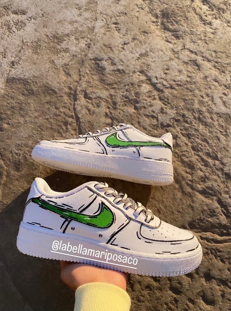 以下が含まれることがあります： 緑と黒の漫画風のナイキのスウッシュが付いた白いスニーカー。靴は黒い輪郭で、明るい茶色の表面の上にあります。靴の下部に「@labellamarposaco」というテキストがあります。