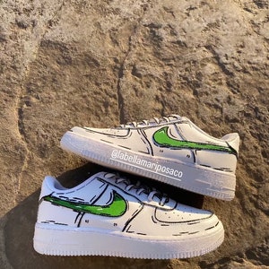 以下が含まれることがあります： 緑と黒の漫画のデザインが施された白いスニーカー。スニーカーには黒いスウッシュロゴがあり、側面には「@labellamarposaco」という文字が書かれています。