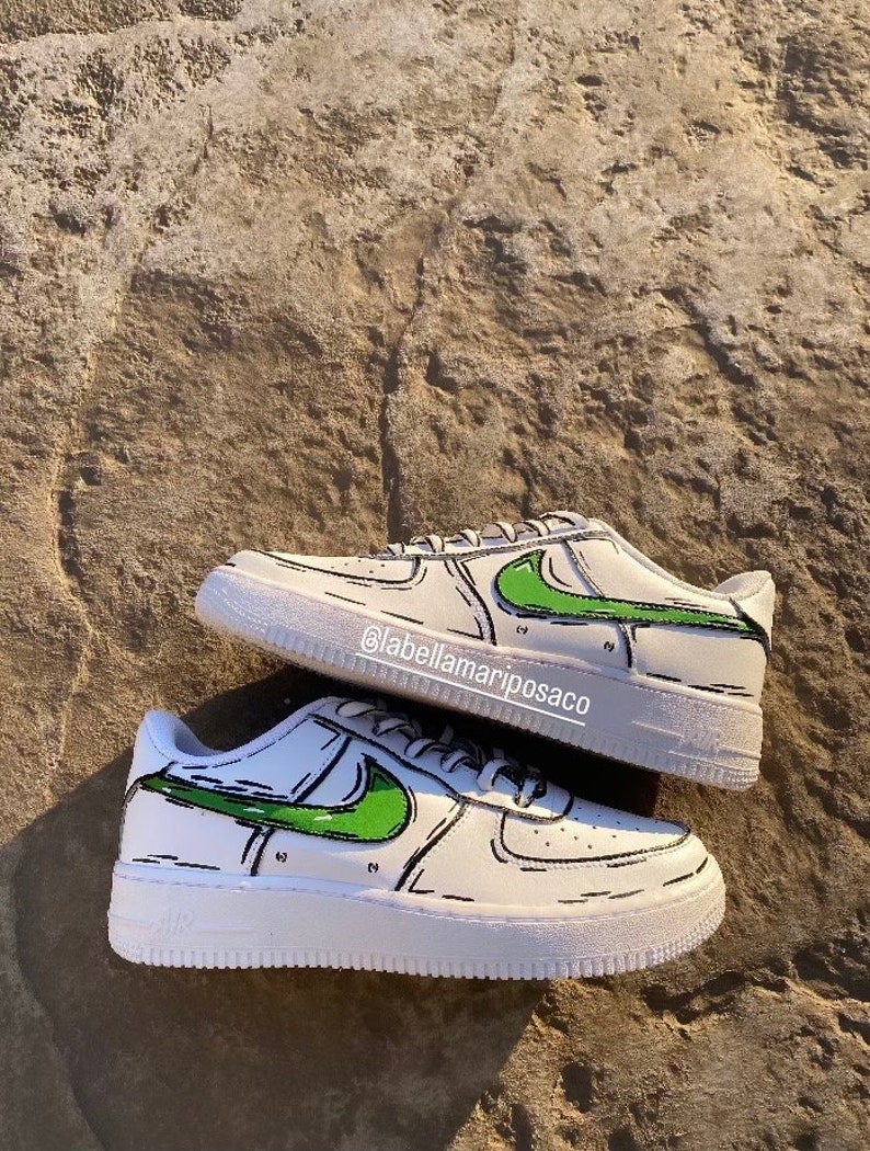 以下が含まれることがあります： 緑と黒の漫画のデザインが施された白いスニーカー。スニーカーには黒いスウッシュロゴがあり、側面には「@labellamarposaco」という文字が書かれています。