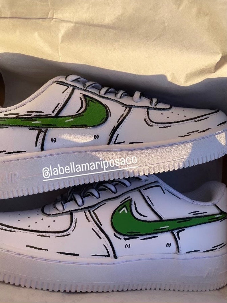 以下が含まれることがあります： 緑と黒の漫画風のナイキのスウッシュがついた白いスニーカー。靴は、側面にInstagramのハンドル@labellamarposacoが書かれた箱に入っています。