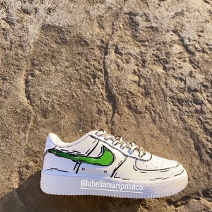 以下が含まれることがあります： 緑と黒の漫画風のナイキのスウッシュが付いた白いスニーカー。靴は白いソールとスウッシュの周りに黒いアウトラインがあります。靴の側面に「@labellamarposaco」というテキストが書かれています。
