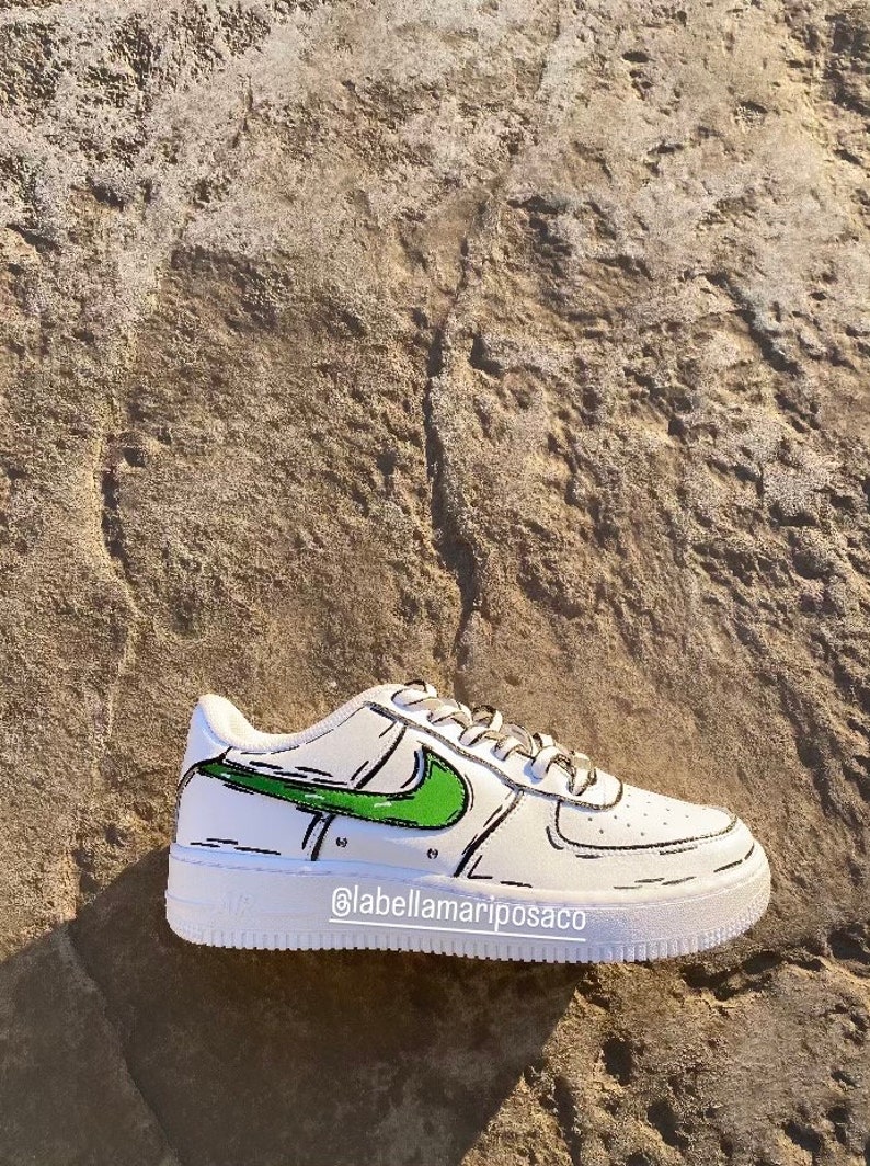以下が含まれることがあります： 緑と黒の漫画風のナイキのスウッシュが付いた白いスニーカー。靴は白いソールとスウッシュの周りに黒いアウトラインがあります。靴の側面に「@labellamarposaco」というテキストが書かれています。