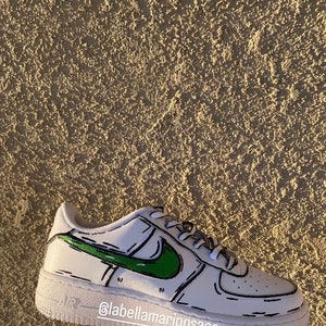 以下が含まれることがあります： 緑と黒の漫画風のナイキのスウッシュが付いた白いローカットスニーカー。靴は白いソールと靴全体を囲む黒いアウトラインを持っています。靴の側面に「@labellamarjposaco」というテキストが書かれています。