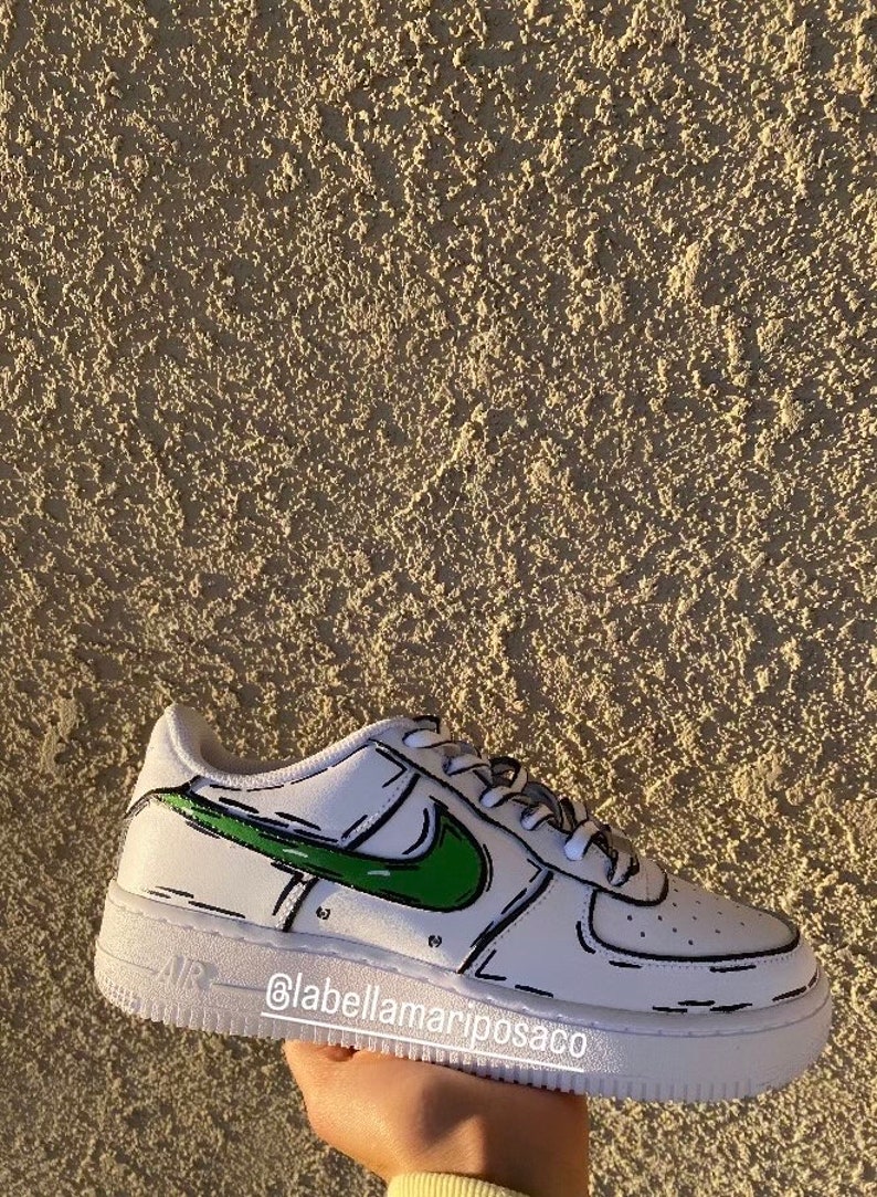 以下が含まれることがあります： 緑と黒の漫画風のナイキのスウッシュが付いた白いローカットスニーカー。靴は白いソールと靴全体を囲む黒いアウトラインを持っています。靴の側面に「@labellamarjposaco」というテキストが書かれています。