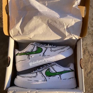 以下が含まれることがあります： 緑色の漫画風のナイキのスウッシュと黒いアウトラインが付いた白いスニーカー。靴は白い箱の中にティッシュペーパーと一緒にあります。