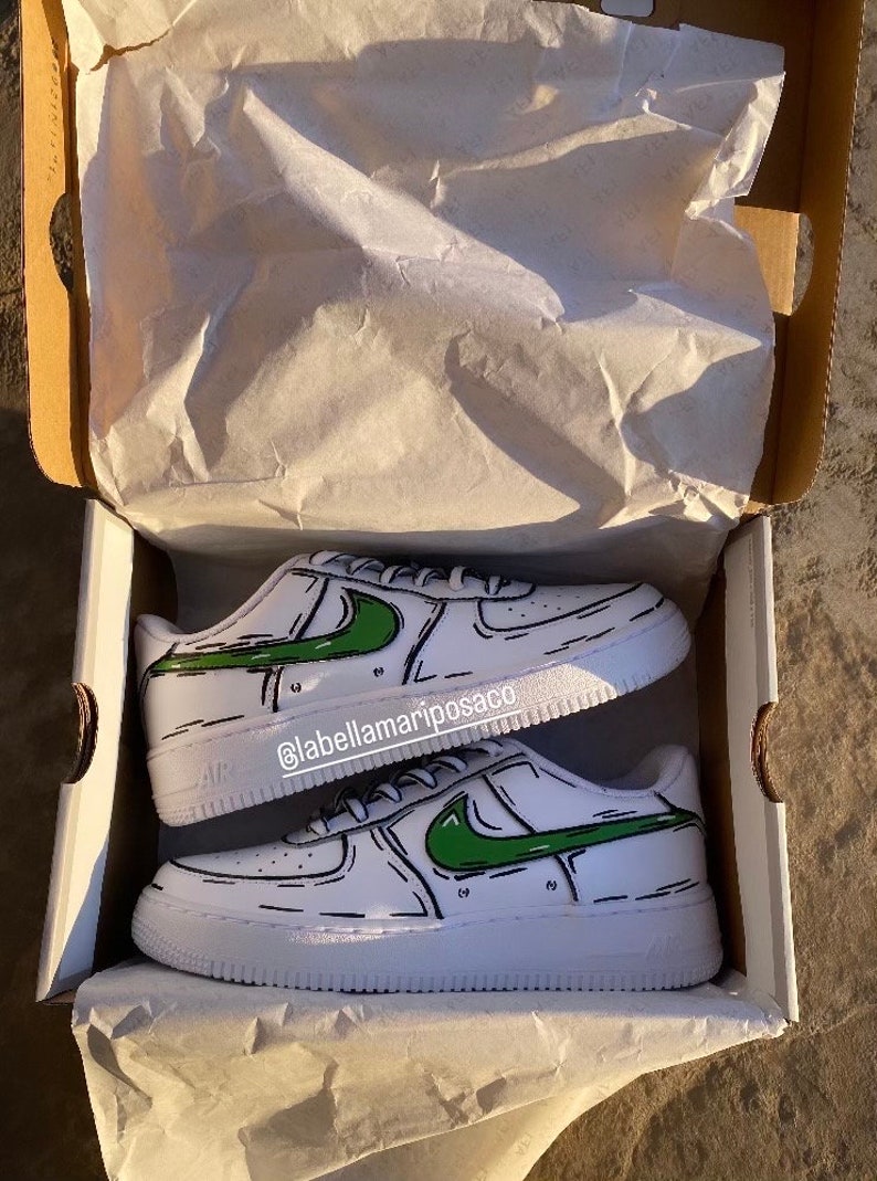 以下が含まれることがあります： 緑色の漫画風のナイキのスウッシュと黒いアウトラインが付いた白いスニーカー。靴は白い箱の中にティッシュペーパーと一緒にあります。