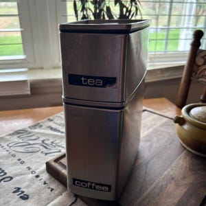 May include: A silver metal canister with a black label that says "tea" on the top and a black label that says "coffee" on the bottom.