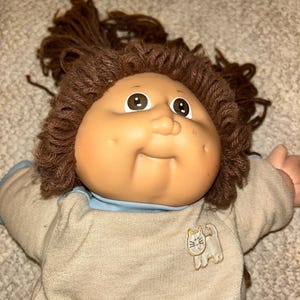May include: A vintage Cabbage Patch Kid doll with brown yarn hair and a tan sweater. The doll has a round face, large eyes, and a small embroidered cat on the sweater. The doll's sweater has blue trim.