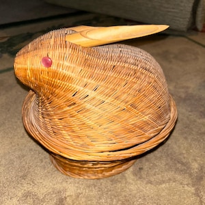 May include: A woven basket shaped like a rabbit, featuring a long, wooden ear and a red bead eye. The basket is made of light brown wicker and has a round base. The rabbit basket is a decorative item.