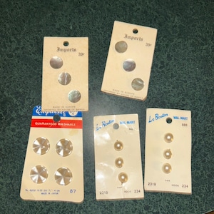 May include: Five vintage retail cards containing sewing buttons. Two cards labeled Imports feature three iridescent shell-like buttons each. One card labeled Exquisite holds four clear faceted buttons. Two Le Bouton cards each hold three small round off-white buttons.