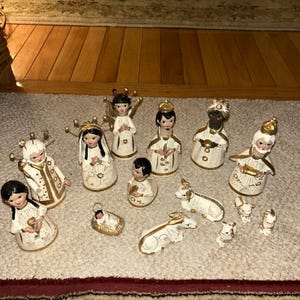 Puede incluir: Una colección de figuras de cerámica del nacimiento. Las figuras son principalmente blancas con detalles dorados y representan ángeles, los tres reyes magos y animales. Las figuras están dispuestas sobre una alfombra de color claro.