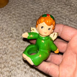 Peut inclure: Petite figurine d'ange en céramique, cheveux roux et tenue verte. L'ange a de petites ailes dorées et est orné d'étoiles jaunes. La figurine est tenue dans une main.