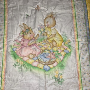 May include: A white quilted blanket featuring a printed illustration of two rabbits having a picnic on a colorful patchwork mat. One rabbit wears a pink dress and floral crown, while the other wears a yellow top. A basket of carrots sits nearby.