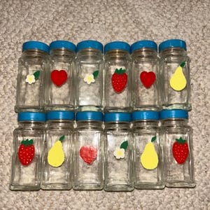 May include: A collection of twelve clear glass spice jars with blue lids. Each jar features a different fruit or flower design, including strawberries, pears, hearts, and flowers. The jars are arranged in three rows of four.