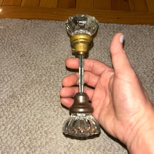 May include: A vintage-style door knob set featuring two clear, faceted glass knobs with a fluted design. The knobs are connected by a central metal spindle and include circular brass-toned and dark metallic mounting plates at the base of each knob.
