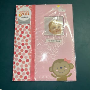 May include: A pink memory book by Just One You made by Carter's, featuring a white vertical stripe with pink polka dots, a square photo window, a label reading my baby book, and a cartoon monkey illustration with a pink bow on its head.
