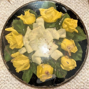 Puede incluir: Bandeja decorativa redonda y negra con un diseño floral. La bandeja presenta flores de papel amarillas y blancas con hojas verdes. Las flores están dispuestas en un patrón circular. La bandeja tiene una apariencia vintage y desgastada.