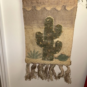May include: A rectangular wall hanging made of woven tan burlap featuring a central tufted green cactus design. The piece includes a smaller green succulent and a rounded plant at the base, finished with a wavy top border and long fringe.