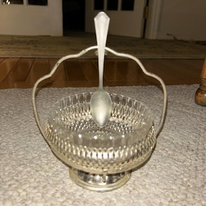 Op de afbeelding: Een zilveren suikerpot met een heldere glazen inzet en een bijpassende lepel. De kom heeft een decoratief handvat en staat op een ronde voet. De glazen inzet heeft een ruitpatroon.