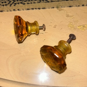 May include: Two amber-colored glass cabinet knobs with metal screws. The knobs have a faceted design and a vintage aesthetic. They are likely used for furniture restoration or as decorative hardware.