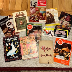 Puede incluir: Una colección de libros de cocina antiguos con cubiertas coloridas. Los títulos incluyen "Ground Beef Cookbook", "Morsels Magic", "Carstairs Party Book" y "Rumford Cook Book". Los libros están dispuestos sobre una superficie clara.