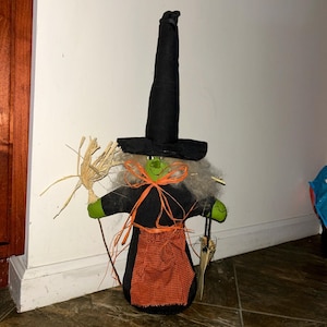 May include: A fabric witch doll featuring a tall black pointed hat, a green face, and gray hair. The doll wears a black dress with an orange and black checkered apron, tied with orange raffia. It holds a small straw broom in one hand.
