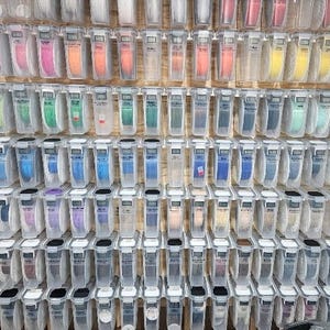 May include: A wall-mounted display featuring numerous clear plastic bins, each containing a spool of ribbon in a distinct color. The grid arrangement showcases a wide spectrum of hues, including reds, oranges, yellows, greens, blues, purples, and neutrals, organized in rows and columns.
