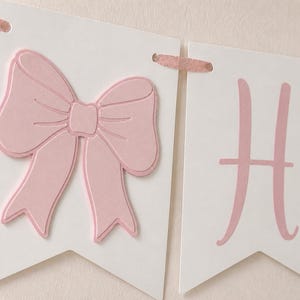 May include: A banner with white pennants and pink accents. One pennant features a large, raised pink bow. Another displays the letter "H" in pink. The banner is strung with pink ribbon.