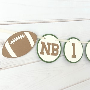 Op de afbeelding: Een banner met een American football-vormige uitsparing en cirkelvormige uitsparingen met de letters "NB" en de cijfers 1, 2 en 3. De uitsparingen hebben groene en crèmekleurige accenten, geregen op een crèmekleurig lint.