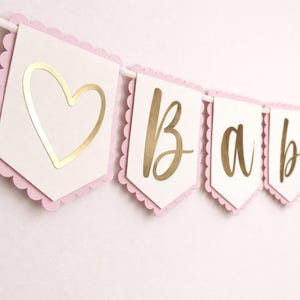 Op de afbeelding: Een babybanner met witte vlaggetjes, elk met een roze geschulpte rand. Gouden letters spellen "BABY" over de vlaggetjes. De eerste vlaggetje heeft een gouden hart.