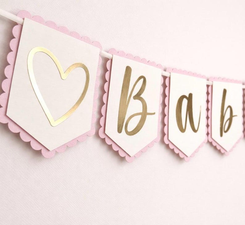 Op de afbeelding: Een babybanner met witte vlaggetjes, elk met een roze geschulpte rand. Gouden letters spellen "BABY" over de vlaggetjes. De eerste vlaggetje heeft een gouden hart.