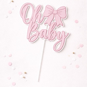 May include: A pink "Oh Baby" cake topper with a bow design. The text is in a cursive font, and the topper is on a white stick. Pink and gold confetti dots the white background.