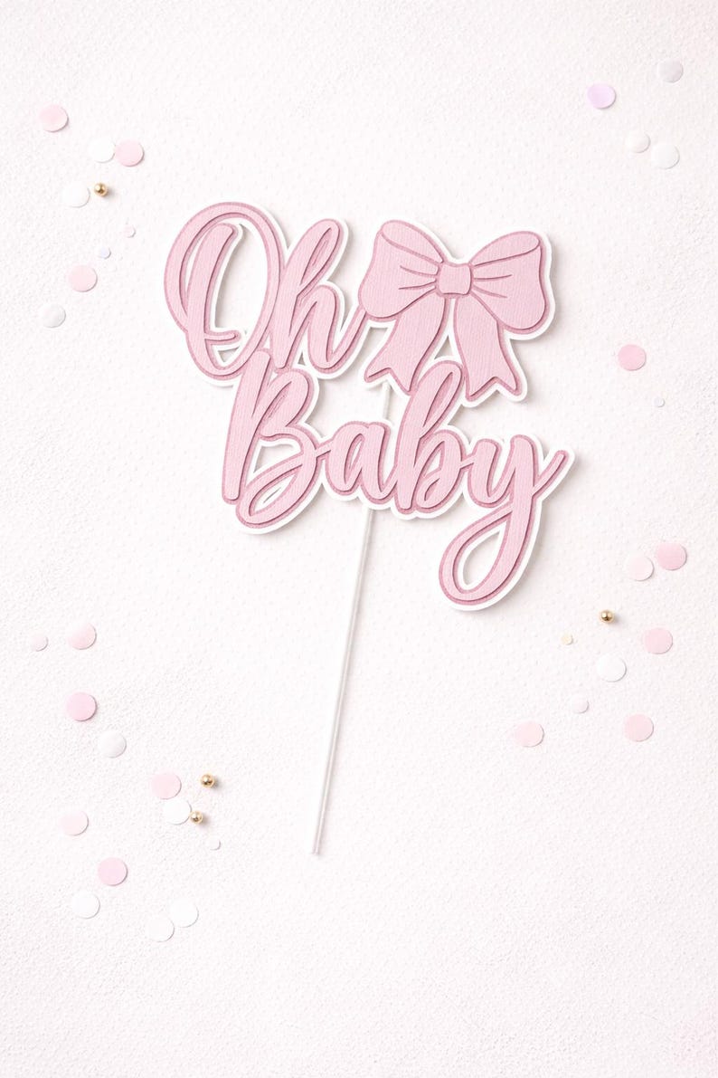 May include: A pink "Oh Baby" cake topper with a bow design. The text is in a cursive font, and the topper is on a white stick. Pink and gold confetti dots the white background.