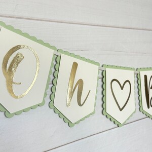 Op de afbeelding: Een slinger met de gouden tekst "Oh Baby". De letters staan op witte vlaggetjes met lichtgroene randen en zijn aan een wit lint geregen. Geschikt voor een babyshower of een soortgelijk feest.
