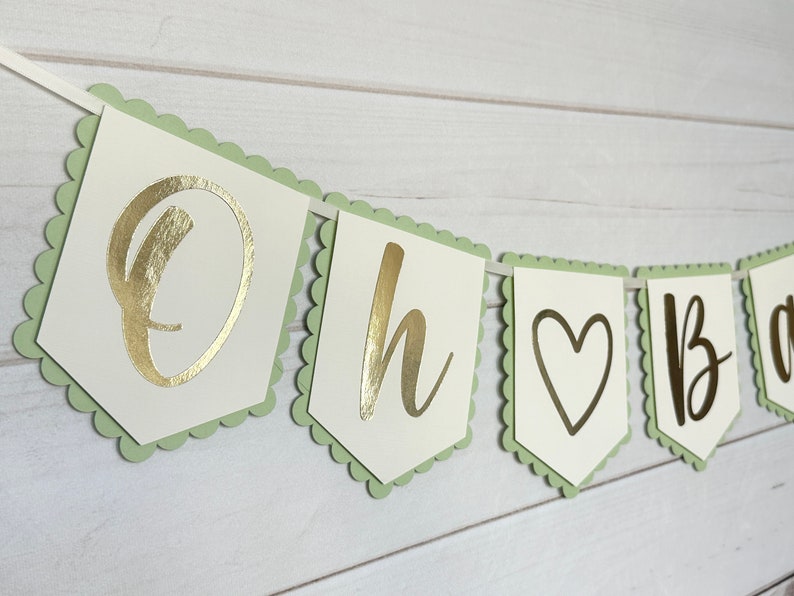 Op de afbeelding: Een slinger met de gouden tekst "Oh Baby". De letters staan op witte vlaggetjes met lichtgroene randen en zijn aan een wit lint geregen. Geschikt voor een babyshower of een soortgelijk feest.
