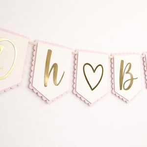Op de afbeelding: Een slinger met de woorden "Oh baby" in gouden letters. De slinger heeft een lichtroze geschulpte rand en is geregen op een wit lint. Het hartsymbool is ook goudkleurig.