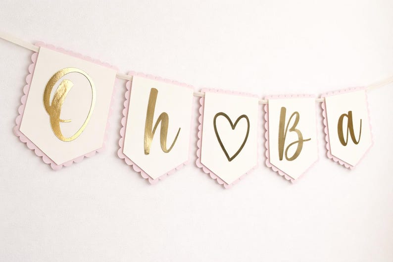 Op de afbeelding: Een slinger met de woorden "Oh baby" in gouden letters. De slinger heeft een lichtroze geschulpte rand en is geregen op een wit lint. Het hartsymbool is ook goudkleurig.