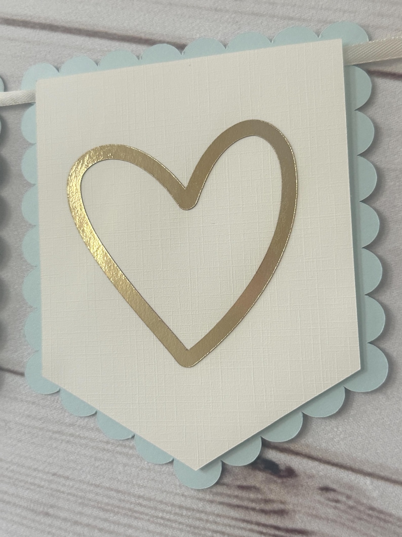 Op de afbeelding: Een banner met een lichtblauwe geschulpte rand en een cr&egrave;mekleurig midden. In het midden van de banner bevindt zich een gouden hartvorm. De banner is geregen op een wit lint.