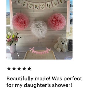 Op de afbeelding: Babyborrel decoratie met een banner "Its a Girl". Roze en witte papieren pompons versieren de achtergrond. Een taart met een roze banner en een teddybeer staan op een tafel. Een vaas met bloemen is ook aanwezig.