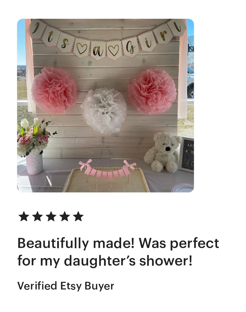 Op de afbeelding: Babyborrel decoratie met een banner "Its a Girl". Roze en witte papieren pompons versieren de achtergrond. Een taart met een roze banner en een teddybeer staan op een tafel. Een vaas met bloemen is ook aanwezig.