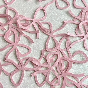 May include: A collection of light pink paper bows scattered across a white, glittery surface. The bows are cut from textured paper and feature a classic bow shape with long, flowing ribbon ends. Ideal for crafting and embellishments.
